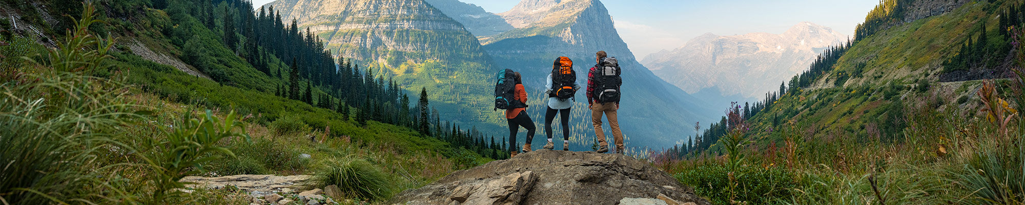 Three friends stands together atop a rocky outcrop wearing their TETON Sports Explorer & Scout backpacks, viewing a large mountain.