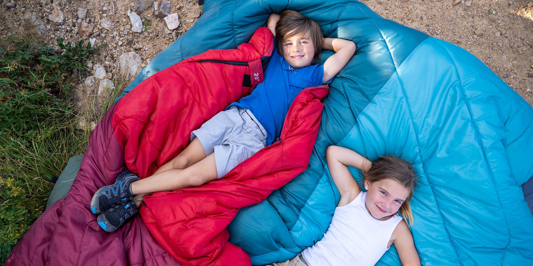 Two children smile up at the camera as they lie together on two brightly colored sleeping bags.