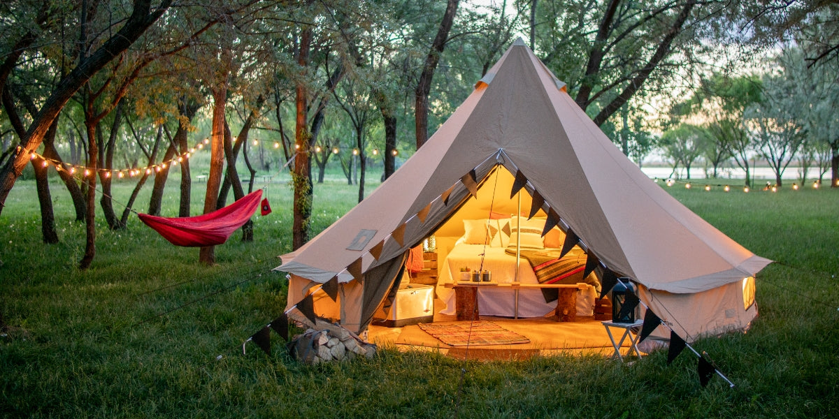 The TETON Sierra Tent: Image shows a bell-shaped canvas tent by TETON Sports setup as a glamping site. There is a large bed in the tent, lights on around and in the tent, a hammock in the background, and just a general serene look and feel.