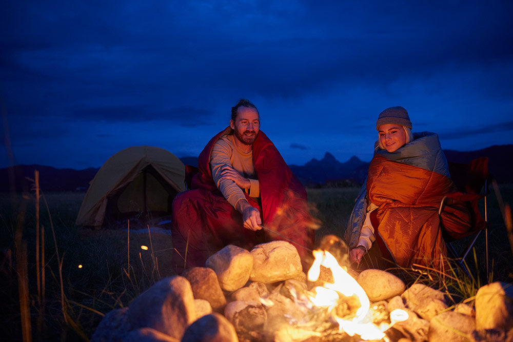 Man and woman roast marshmallows in camp blankets.