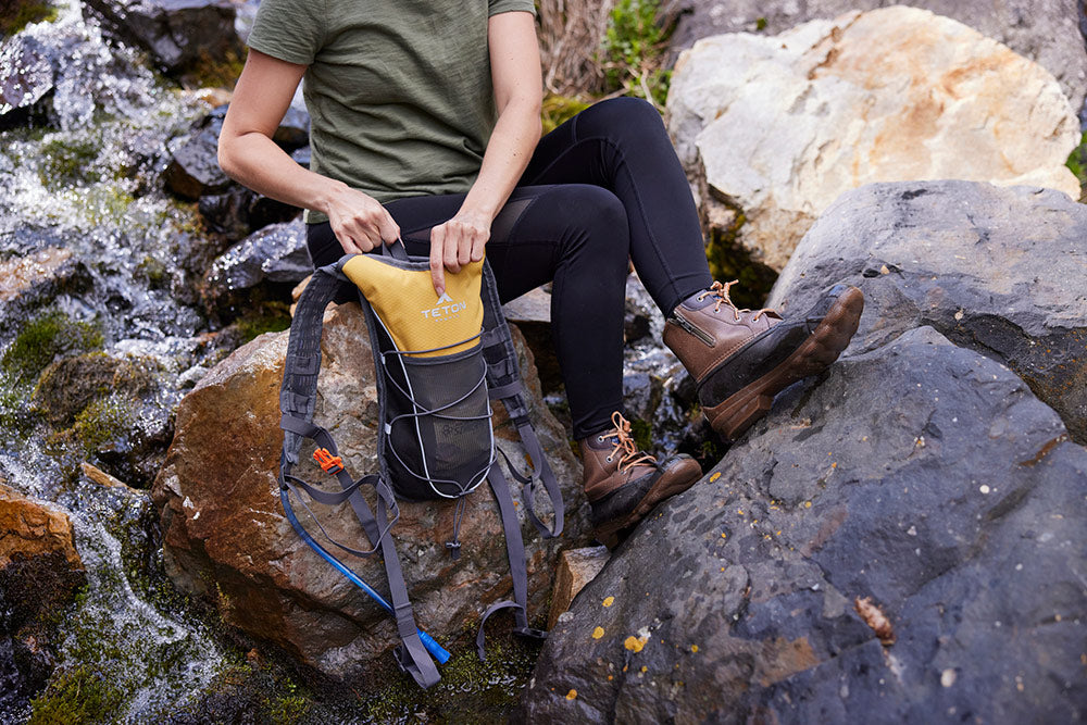 Woman holds hydration pack while sitting on rock.