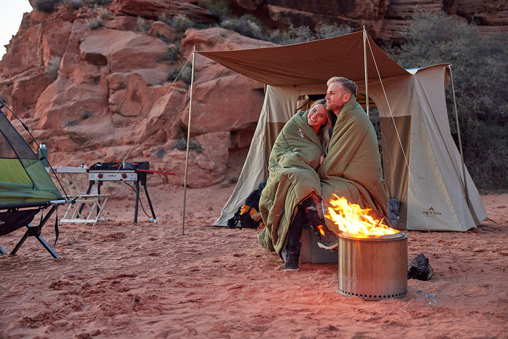 Couple cuddles around fire in sleeping bag while camping.