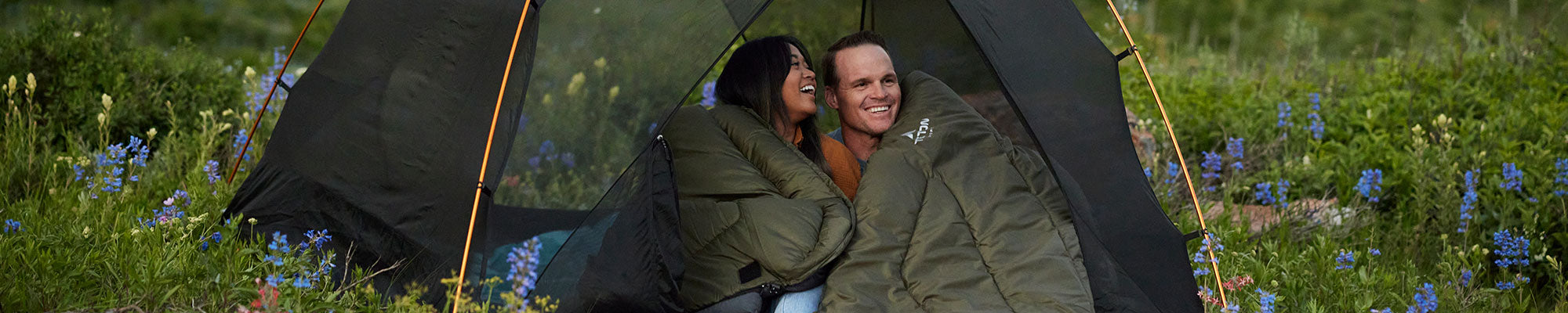 A couple laughs together by a campfire while snuggled up in a TETON Sports Evergreen Mammoth Sleeping Bag.