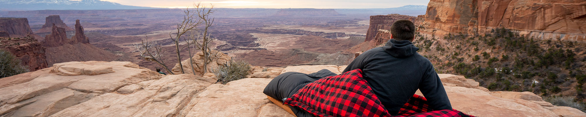 A man sits in his sleeping bag looking out at a scenic view.