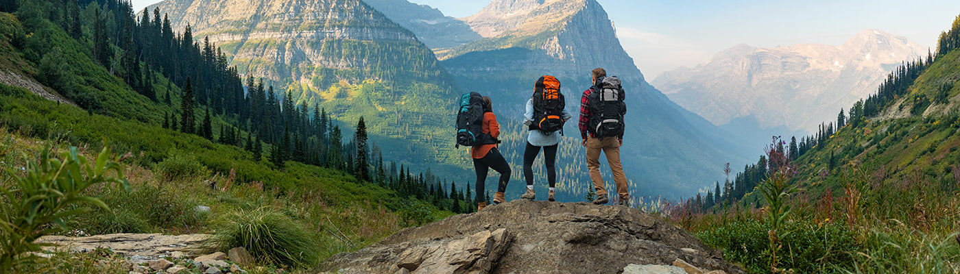 Three friends stands together atop a rocky outcrop wearing their TETON Sports Explorer & Scout backpacks, viewing a large mountain.