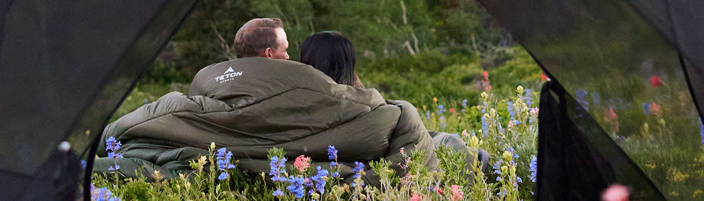 A man and a woman are seen through the open tent doors sitting in a field of wildflowers and snuggling up in their Mammoth sleeping bag.