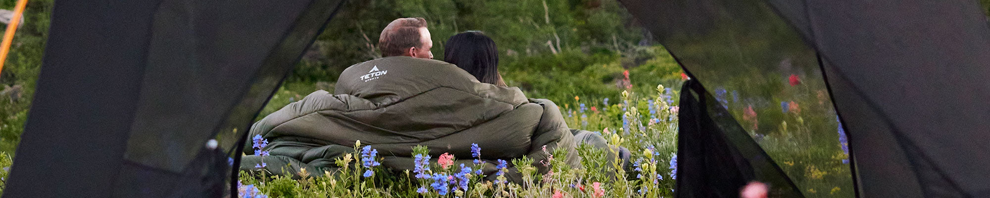 A man and a woman are seen through the open tent doors sitting in a field of wildflowers and snuggling up in their Mammoth sleeping bag.