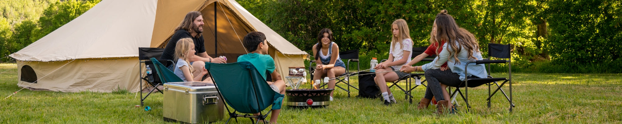 A family camps together around a campfire while their TETON Sports Sierra Canvas Bell Tent sits in the background.