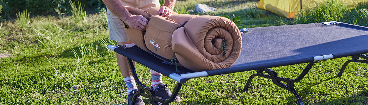A man unbuckles his rolled up TETON Sports canvas camp pad atop his Outfitter XXL camp cot.