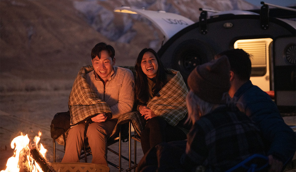 Friends huddled around a campfire just after dusk. There is a man and a woman snuggled together in a TETON Sports Evergreen Mammoth Sleeping Bag and they are facing the camera and smiling. 
