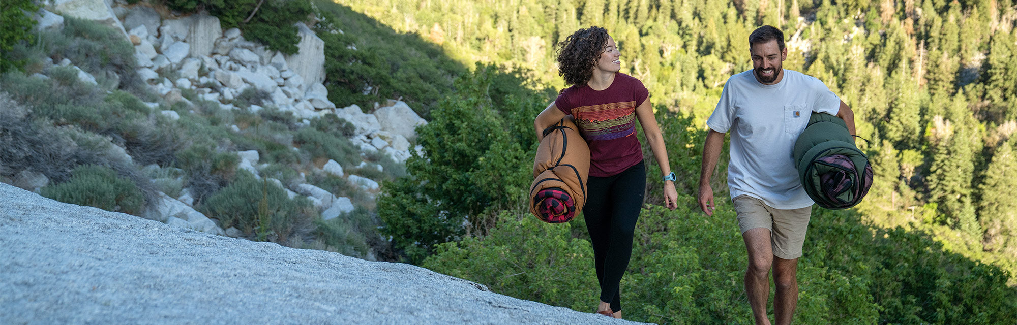 Image shows a couple hiking together along on a granite boulder with a pine forest in the background. They are each carrying TETON Sports Bridger Canvas Sleeping Bags that are rolled for storage.