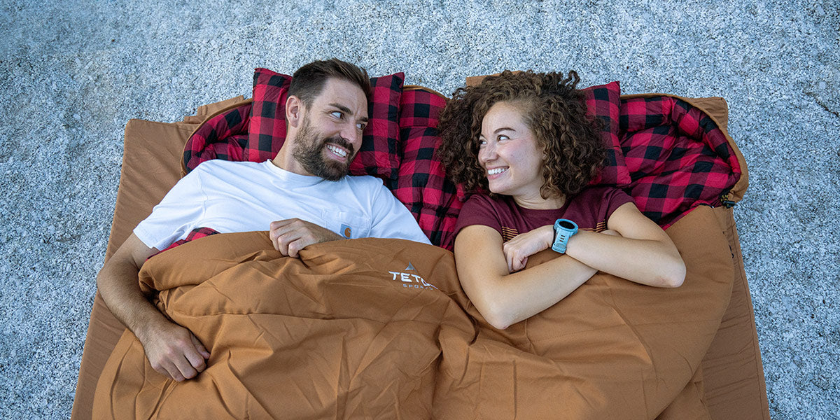 Image shows a couple snuggled up together in a canvas TETON Sports Bridger Mammoth Sleeping Bag in brown. They are looking at each other and smiling. The background is granite.