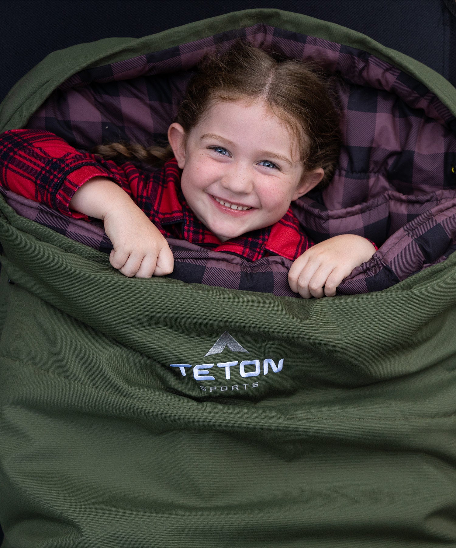 Image shows a young girl smiling while snuggled in a green TETON Sports Bridger Canvas Sleeping Bag.