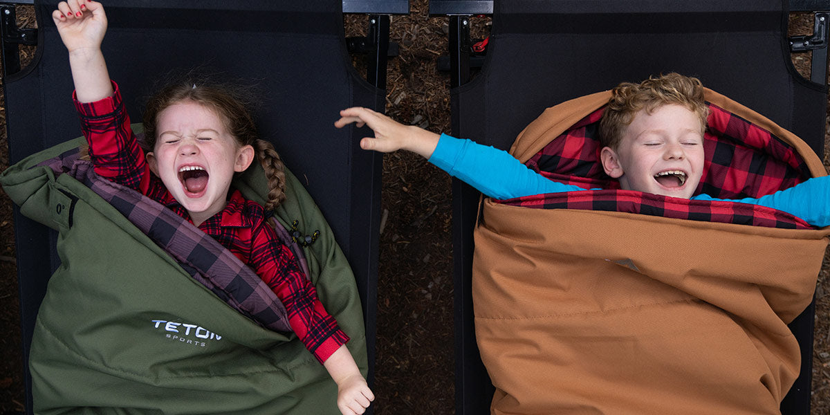 Image shows two children snuggled up in TETON Sports L'il Bridger Sleeping Bags. There is a boy in a brown bag and a girl in a green bag. Both are stretching like they are tired and are sleeping on a TETON Sports Adventurer Cot.
