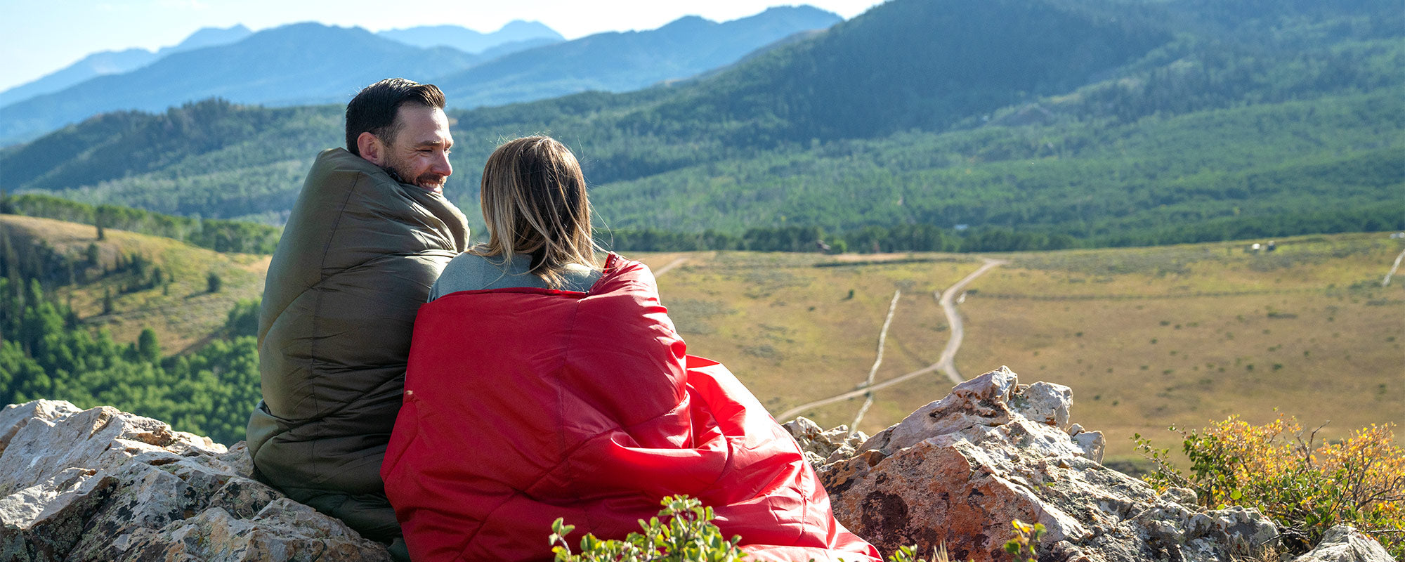 A man and a woman sit together on a rocky outcrop snuggled up in a new Celsius Sleeping Bag by TETON.