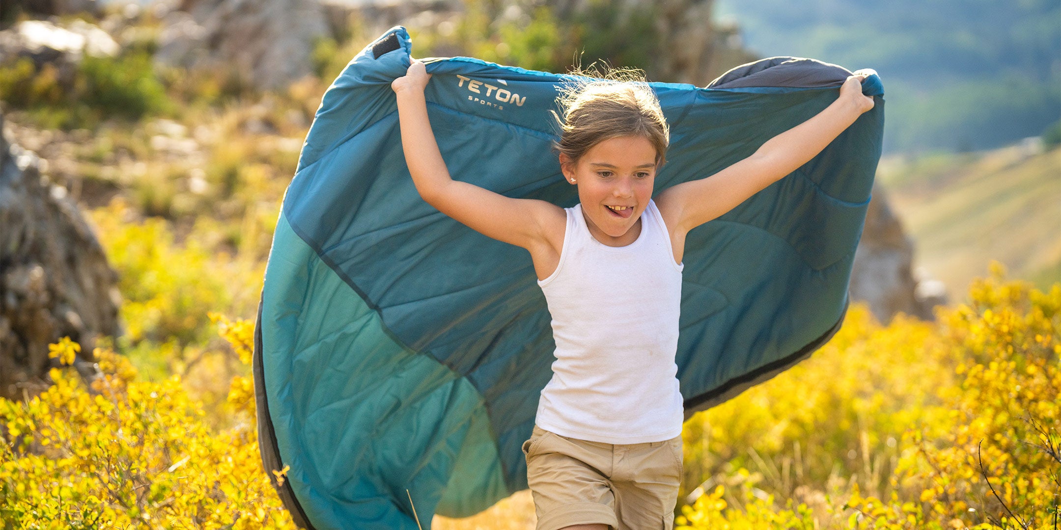 A young girl runs through a field of wildflowers while holding her sleeping bag as a cape.
