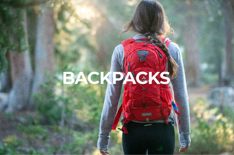 Shop Backpacks: Image shows a female hiking in the woods wearing a TETON Sports Oasis Hydration Backpack in red. She is facing away from the camera.